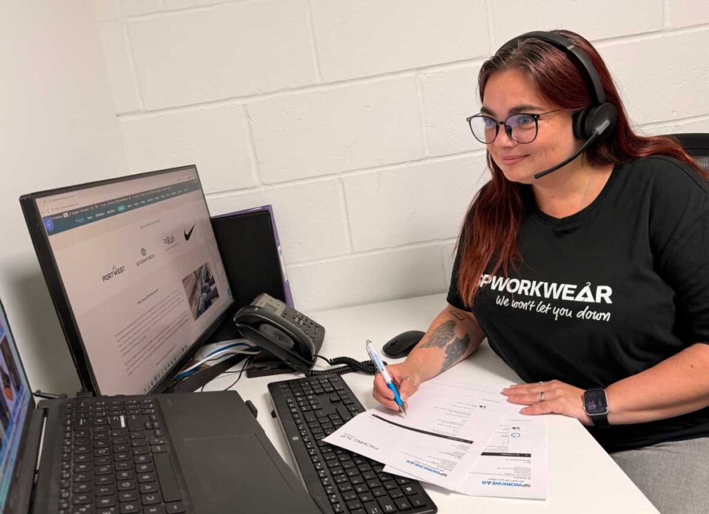Woman on a headset works at a desk with a laptop, monitor, and paperwork.
